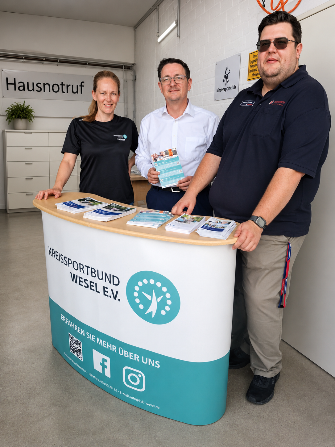 Zwei Männer und eine Frau stehen vor einer Werbetheke mit der Aufschrift "Kreissportbund Wesel".