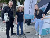 Zwei Mitarbeiterinnen des Kreissportbundes stehen mit einem Mann, der einen Musikkoffer trägt, vor einer Beachflag des Kreissportbund Wesel.