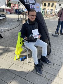Ein junger Mann, sitzend auf einem Stuhl, mit einer Urkunde in der Hand. An Armen, Beinen, Oberkörper und Nacken trägt er Gewichtsmanschetten.