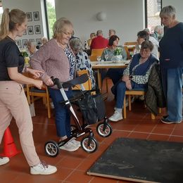 Eine jüngere Frau erklärt einer älteren Dame mit Rollator, wie sie über eine vor ihr auf dem Boden liegende Platte kommt. Im Hintergrund stehen und sitzen weitere ältere Personen und schauen zu.