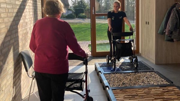 Zwei Personen mit Rollatoren in einem hellen Flur, eine Person steht auf einem taktilen Übungsparcours mit verschiedenen Oberflächen, die andere nähert sich von hinten.
