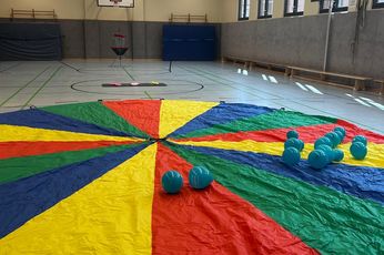 Ein großes, buntes Schwungtuch liegt ausgebreitet in einer Turnhalle. Auf ihr liegen knapp zwei Duzend türkise Luftballons. Im Hintergrund ist das Spiel Discgolf zu sehen.