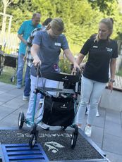 Eine Pflegekraft in grauer Kleidung benutzt einen Rollator, während sie eine Rampe hinuntergeht. Im Hintergrund sind weitere Personen sichtbar.