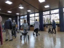 Eine Gruppe Menschen fährt mit Rollatoren durch eine Turnhalle.