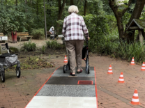 Eine ältere Dame geht mit dem Rollator von der Kamera weg über ein Hindernis aus Kopfsteinpflaster, Fliesen und Rasen. Neben ihr stehen mehrere Pylonen.