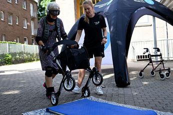 Eine Frau im gemusterten Kleid, mit Gewichtsmanschetten an Ellbogen, Hand-, Knie- und Fußgelenken kippt einen Rollator zum Überqueren einer blauen Turnmatte. Neben ihr steht eine Mitarbeiterin des Kreissportbundes und gibt ihr Tipps.
