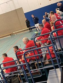 Mehrere Männer in roten Sportjacken sitzen auf einer Tribüne in einer Sporthalle, im Hintergrund stehen zwei Männer, einer in schwarzer Sportkleidung und einer in weißer Hose und dunklem Oberteil.