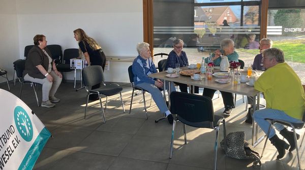 Mehrere ältere Personen sitzen an einem Tisch mit Getränken und Snacks in einem hellen Raum mit großen Fenstern.