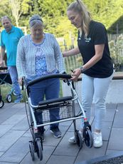 Eine ältere Frau mit einem Rollator wird von einer Mitarbeiterin des KSB unterstützt. Die ältere Frau trägt einen grauen Pullover und ein gemustertes Oberteil. Die jüngere Frau trägt ein schwarzes T-Shirt und eine helle Hose.