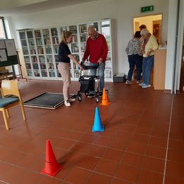 Eine jüngere Frau erklärt einem älteren Herrn einen Rollator. Im Vordergrund stehen Pylonen eines Rollatorparcours.