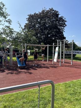 Eine Frau hangelt sich an einer Monkeybar und ein Mann macht Situps an einer weiteren Station, während einige andere Personen zuschauen.