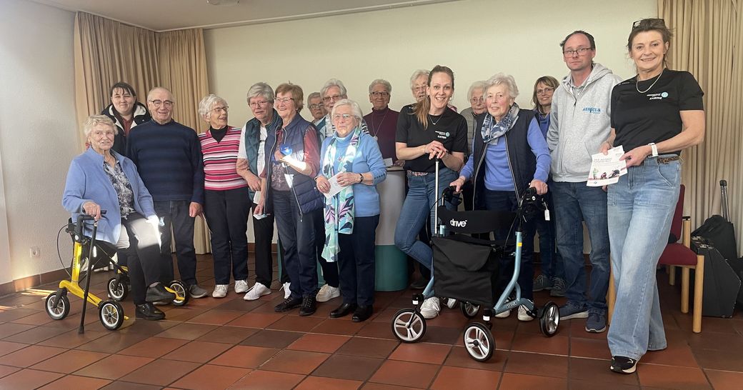 Eine Gruppe Seniorinnen und Senioren, teilweise mit Rollatoren, die Rollatorführerscheine und Präsente in den Händen halten. Zwei Mitarbeiterinnen des Kreissportbundes und der Firma Aericon stehen zwischen den Seniore*innen.