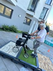 Eine ältere Frau mit einem Rollator steht auf einer Plattform mit unterschiedlichen Oberflächen. Eine Mitarbeiterin des KSB hilft ihr dabei. Im Hintergrund sind Gebäude und Pflanzen sichtbar.