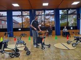Ein Mann steht mit einem Rollator in einer Turnhalle und erklärt. Eine Frau und ein Mann stehen im Hintergrund und hören zu. Im Vordergrund stehen Rollatoren.
