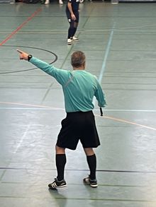 Schiedsrichter steht in einer Halle und zeigt mit dem linken Arm nach links.