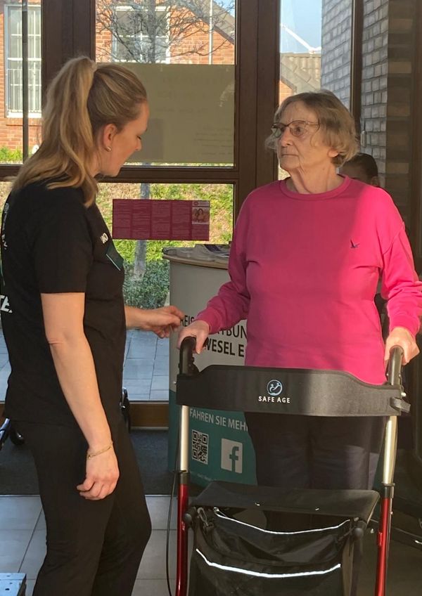 Eine ältere Frau mit Rollator spricht mit einer jüngeren Frau in schwarzer Kleidung vor großen Fenstern in einem hellen Raum.