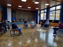 Mehrere Menschen in Rollstühlen stehen in einer Turnhalle.