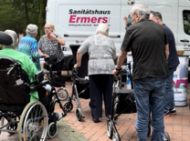 Ein weißer Kleinbus des Sanitätshauses Ermers, vor dem einige Seniorinnen und Senioren mit ihren Rollatoren und Rollstühlen stehen.